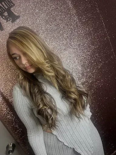 A person with long, layered, wavy blonde hair wears a light grey, ribbed, long-sleeved top against a dark, glittery wall.