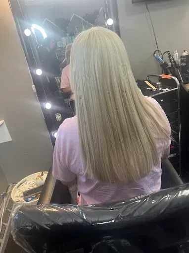 A person sits in a salon chair facing a mirror, showcasing long, straight, light-blonde hair.