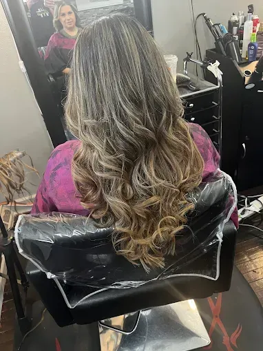 A person in a salon chair with long, wavy, highlighted brown hair, reflected in a mirror.