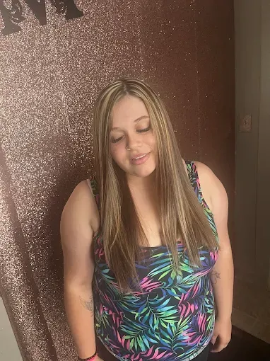A person with long, highlighted brown hair looking down, wearing a sleeveless, tropical-patterned top against a glittery wall.