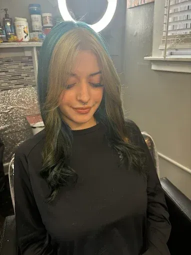 A person with long hair featuring blonde face-framing highlights and dark teal-toned ends, sitting in a salon setting.