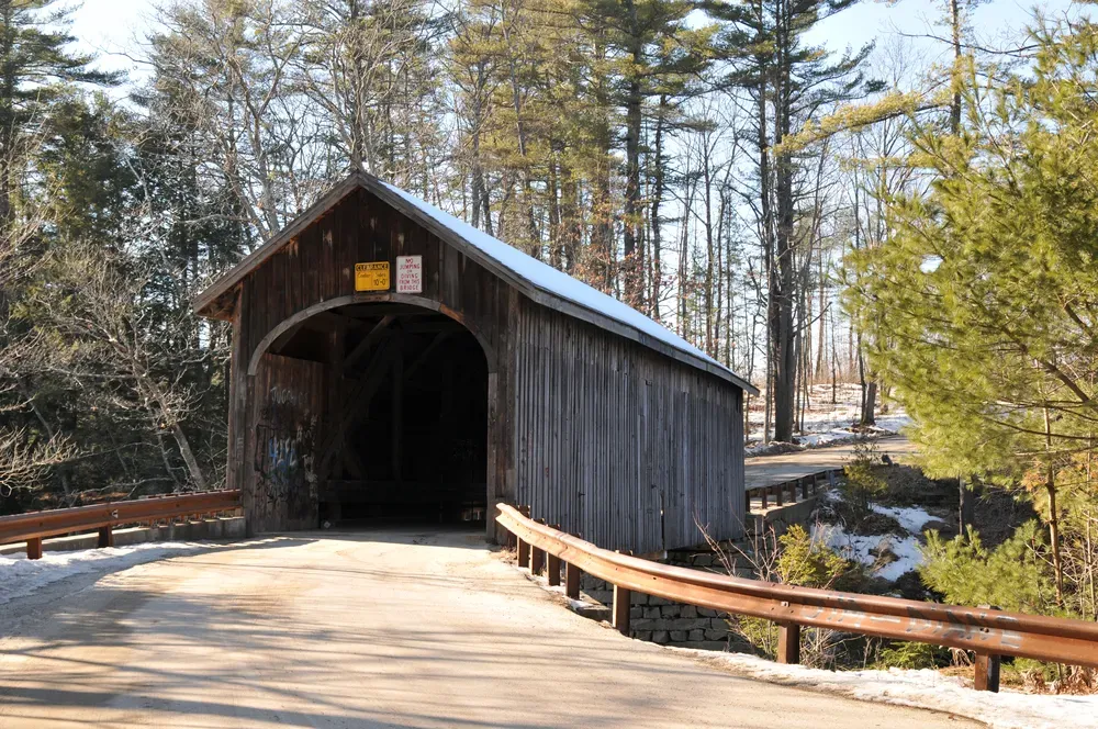 Covered wooden bridge in a forest, with a curved guardrail and sunlight on the road.