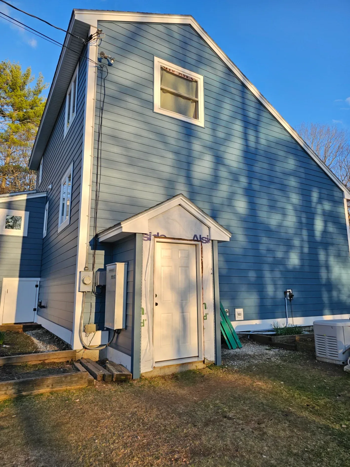 Blue two-story house exterior with a small shed-like entryway and a window, lit by late-afternoon sun.