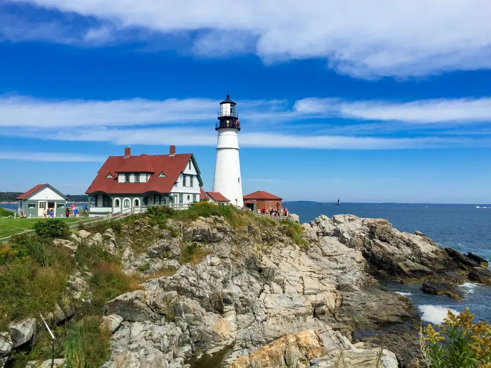 White lighthouse beside red-roofed house on rocky coastline under a blue sky