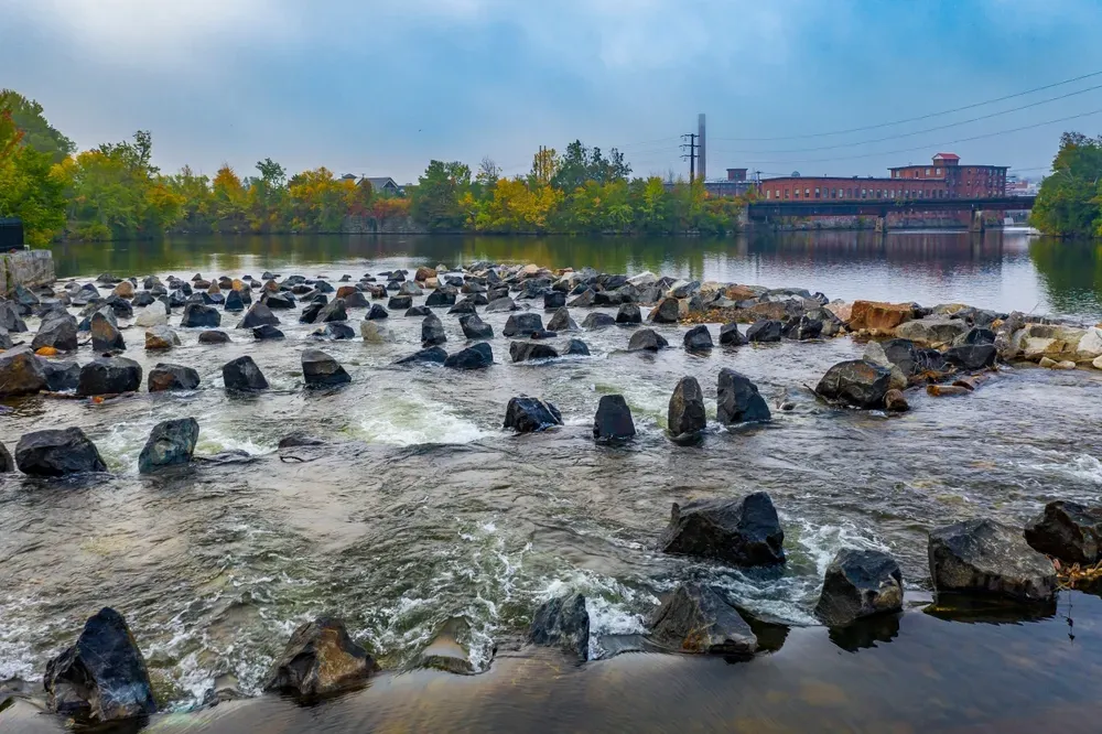 Rushing river with many rocks in the foreground, trees along the shore, and buildings under a blue sky.