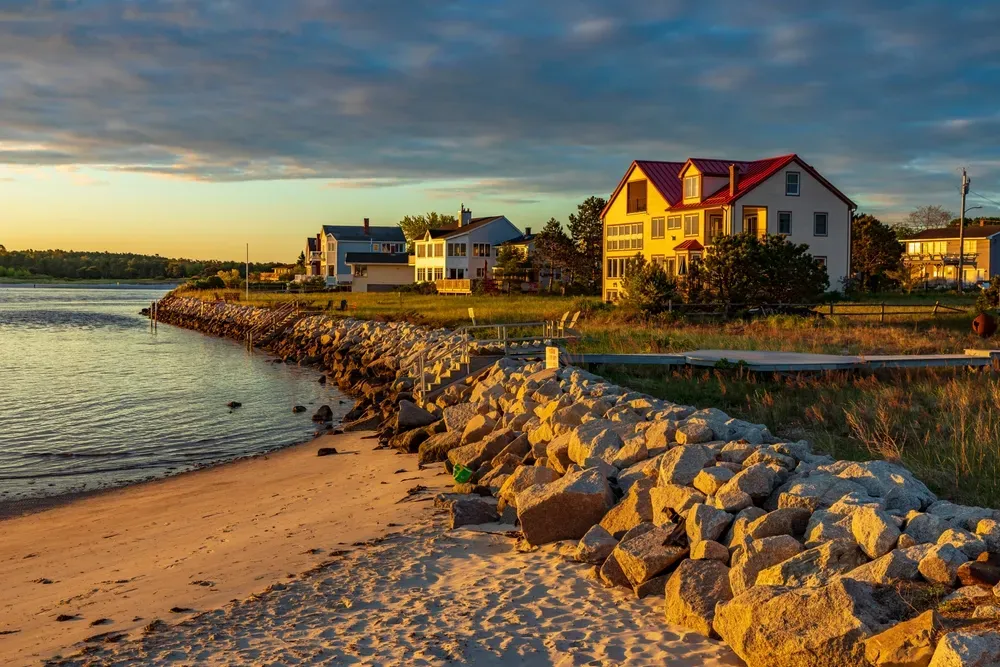 Rocky shoreline with beach houses at sunset, warm light on the water and sandy shore