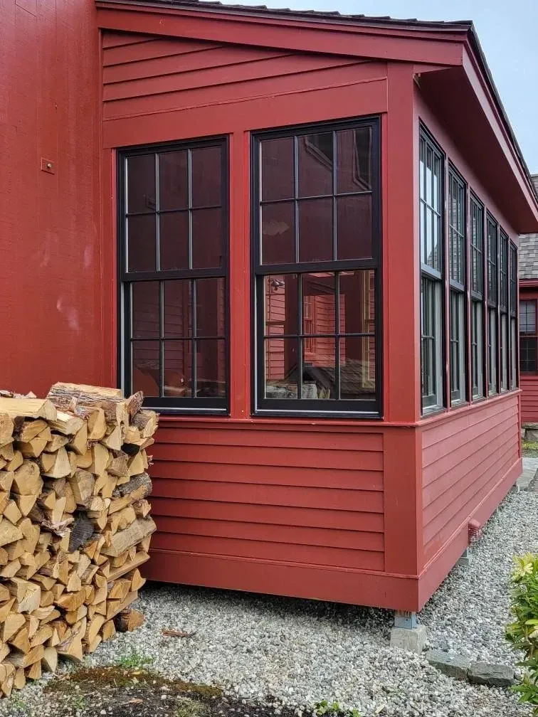 Red house exterior with black-framed windows, a woodpile stacked beside it, and gravel ground.