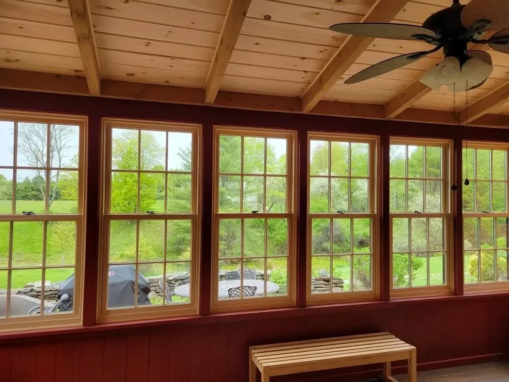 Wood-paneled sunroom with large windows, a ceiling fan, and a view of trees and parked cars outside.
