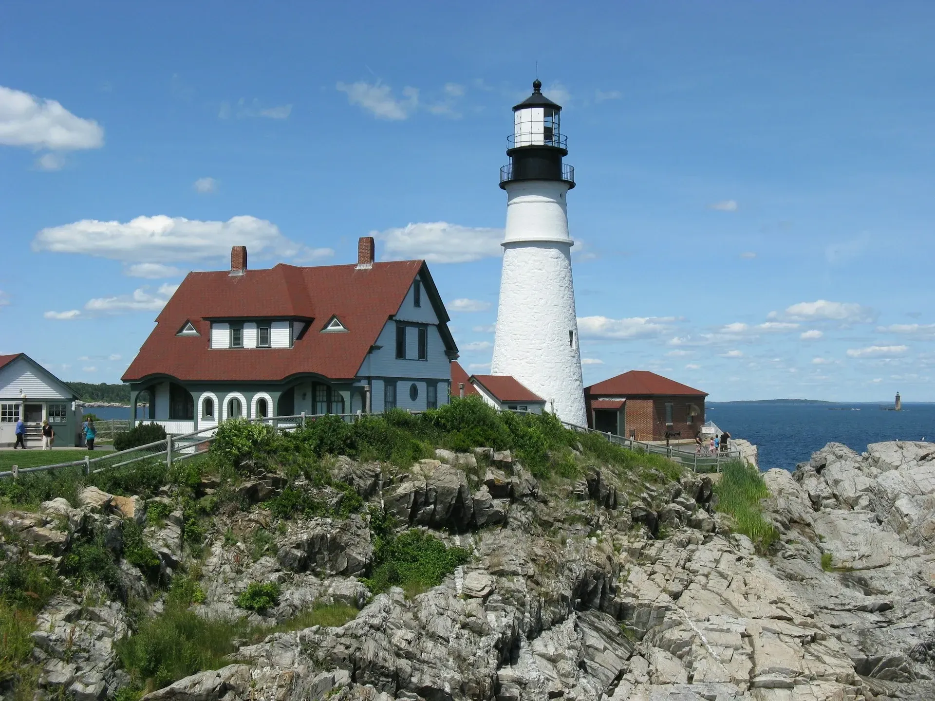 White lighthouse beside red-roofed house on a rocky coastline under a blue sky