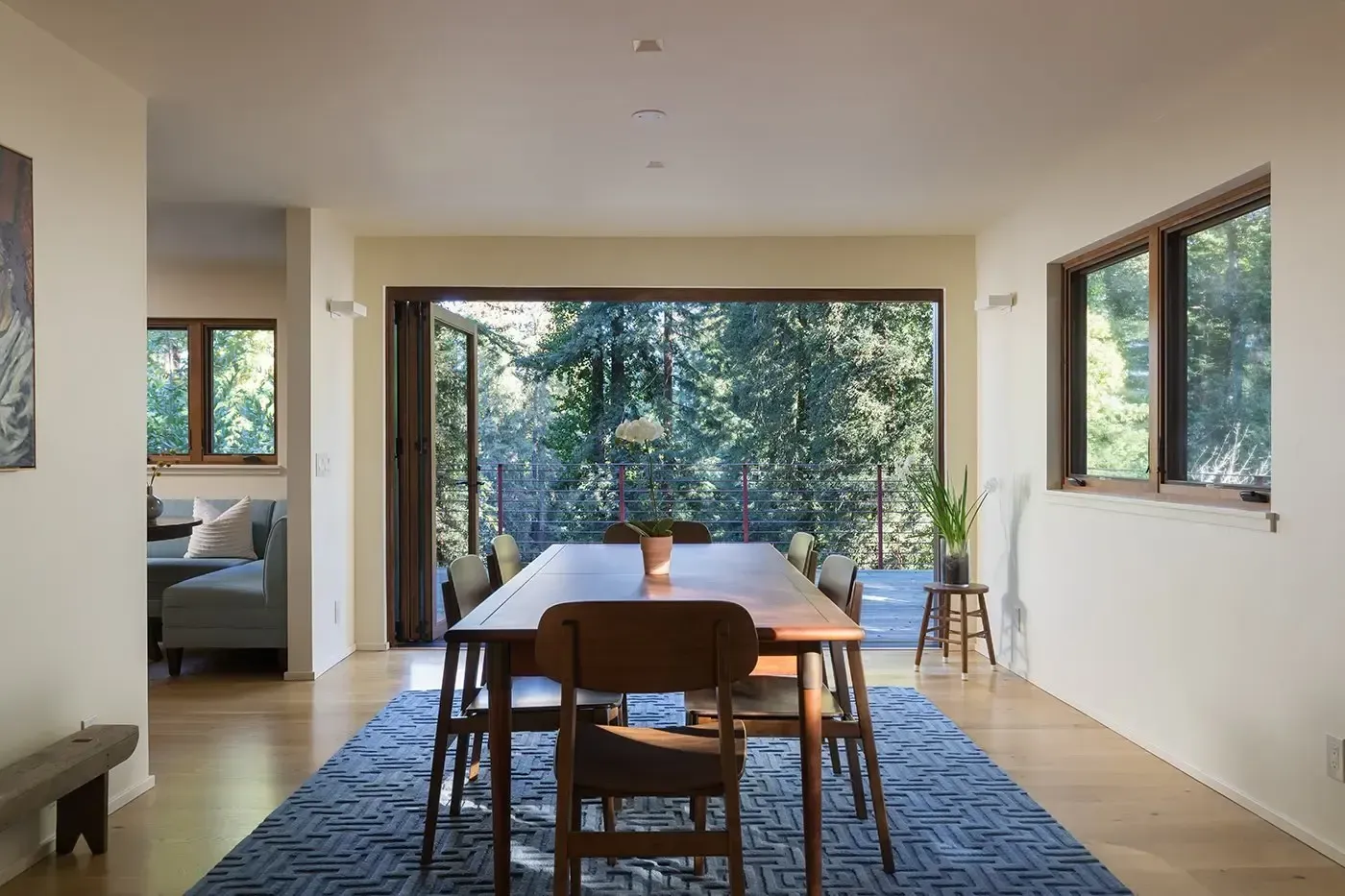 Modern dining room with a wooden table, blue rug, and large windows overlooking trees