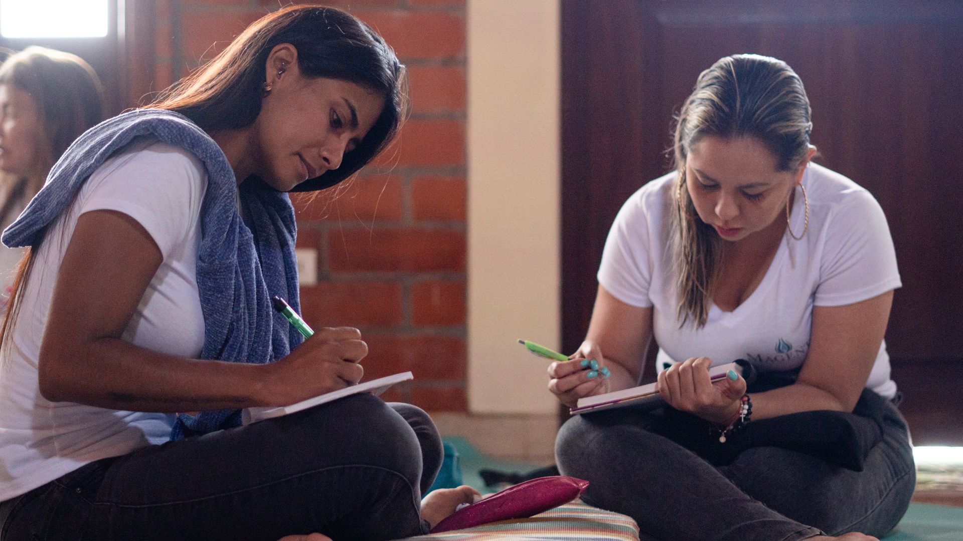 Dos mujeres sentadas en el suelo escribiendo en cuadernos, luz del sol, pared de ladrillos.