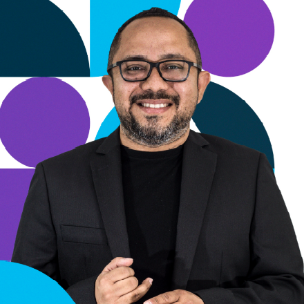 Hombre con gafas y barba, sonriendo, con blazer negro. Formas geométricas abstractas de fondo.