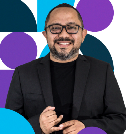 Hombre con gafas, blazer negro y sonriente, sobre un fondo geométrico de formas azules y violetas.