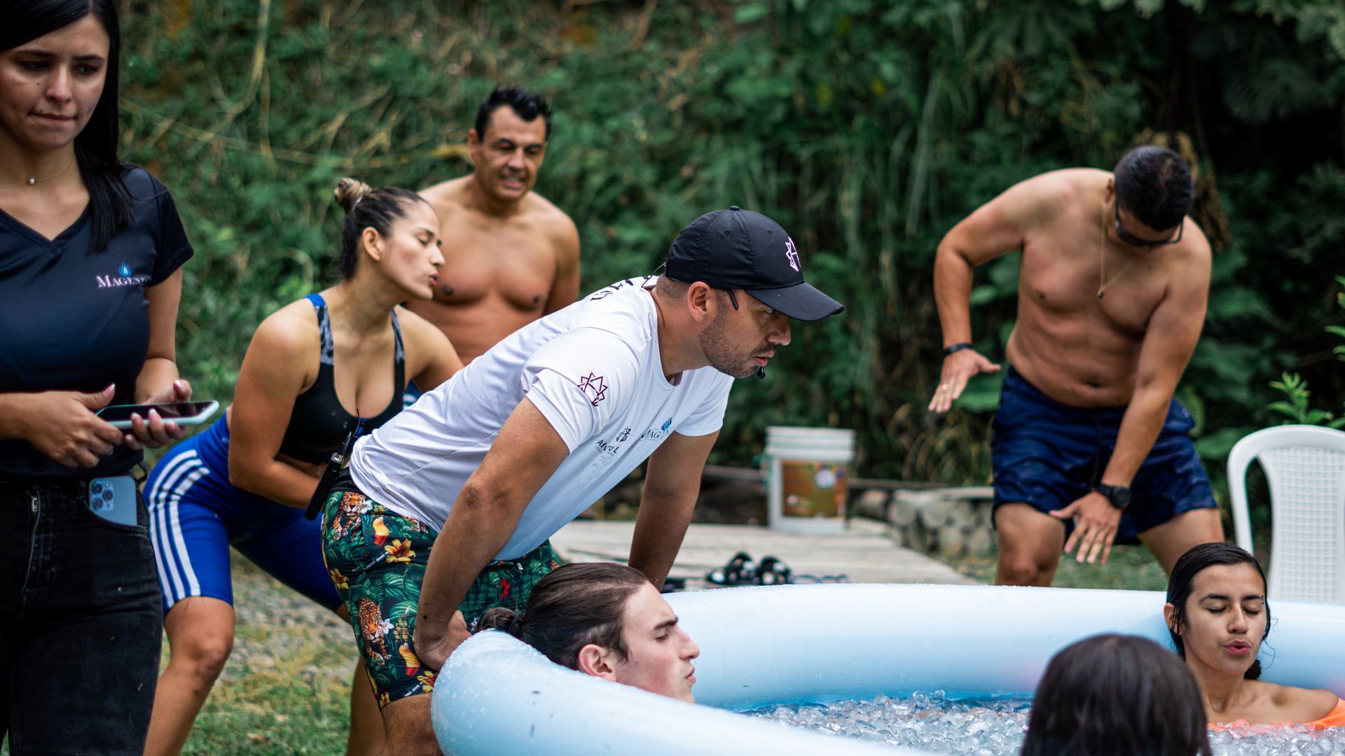 Personas dentro y alrededor de una pequeña piscina, una dando instrucciones. Al aire libre, algunos sin camisa, otros con ropa deportiva.