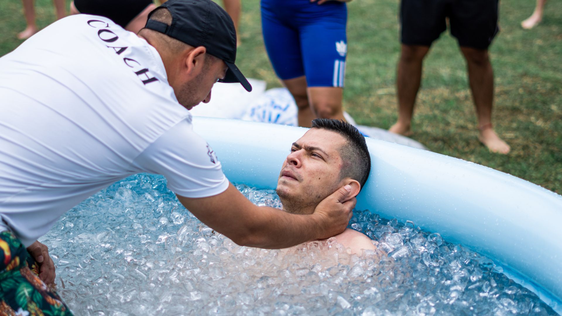 Hombre sumergido en baño de hielo; entrenador sujetándose la cabeza, al aire libre.