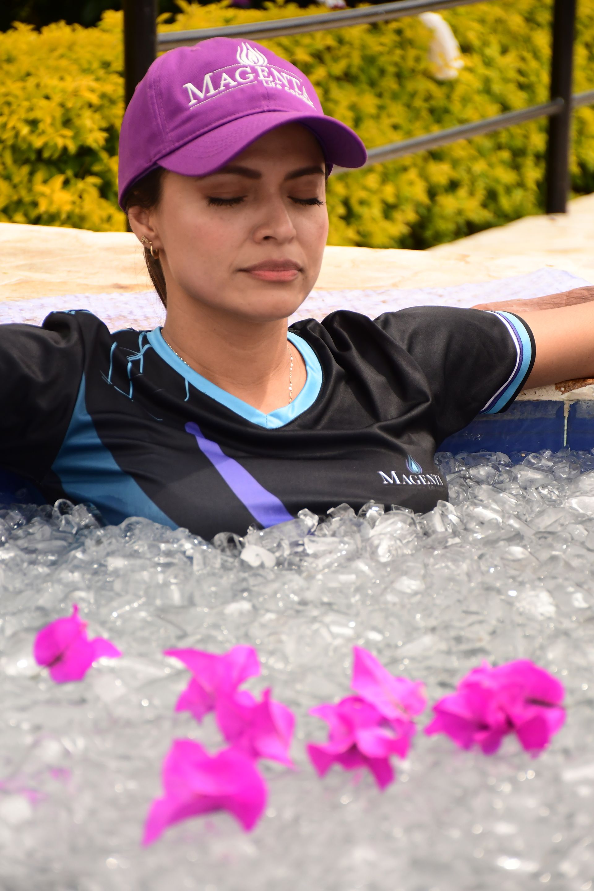 Mujer con gorra morada y camisa oscura relajándose en una piscina, con los ojos cerrados, con flores rosas.