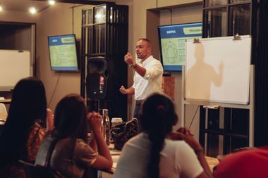 Hombre dando una presentación a una audiencia sentada, señalando una pizarra y pantallas en una sala.