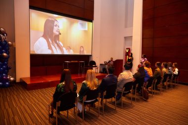 El público observa a una mujer en una pantalla realizando una presentación en un espacio de eventos moderno.