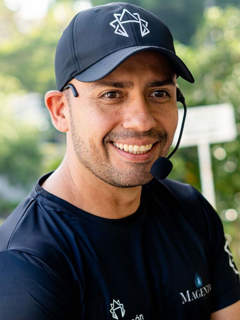 Hombre sonriendo, vistiendo una gorra negra con logo y micrófono, al aire libre.