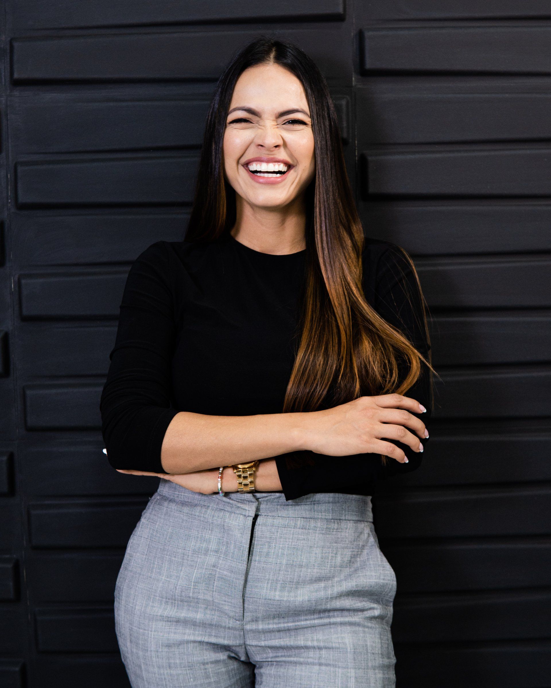 Mujer sonriendo, vestida con un top negro y pantalones grises, apoyada contra una pared negra texturizada.