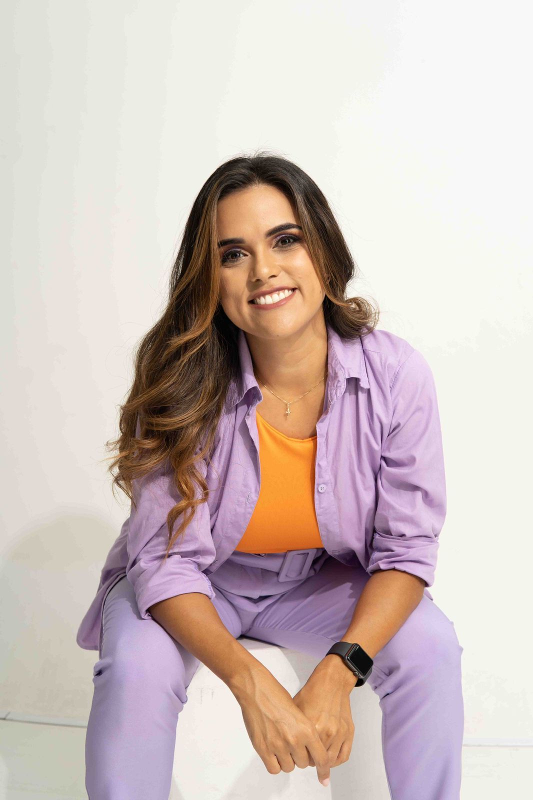 Mujer sentada, sonriente, vestida con una camiseta naranja, una camisa morada y pantalones, con cabello oscuro, sobre un fondo blanco.