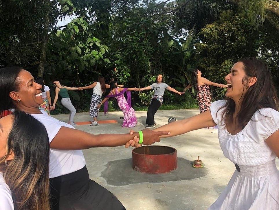 Grupo de personas tomadas de la mano y riendo en un círculo de yoga al aire libre, en un entorno verde y natural.