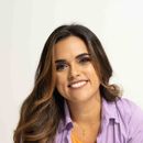 Mujer de cabello castaño, sonriente, vistiendo una camisa morada sobre un top naranja.