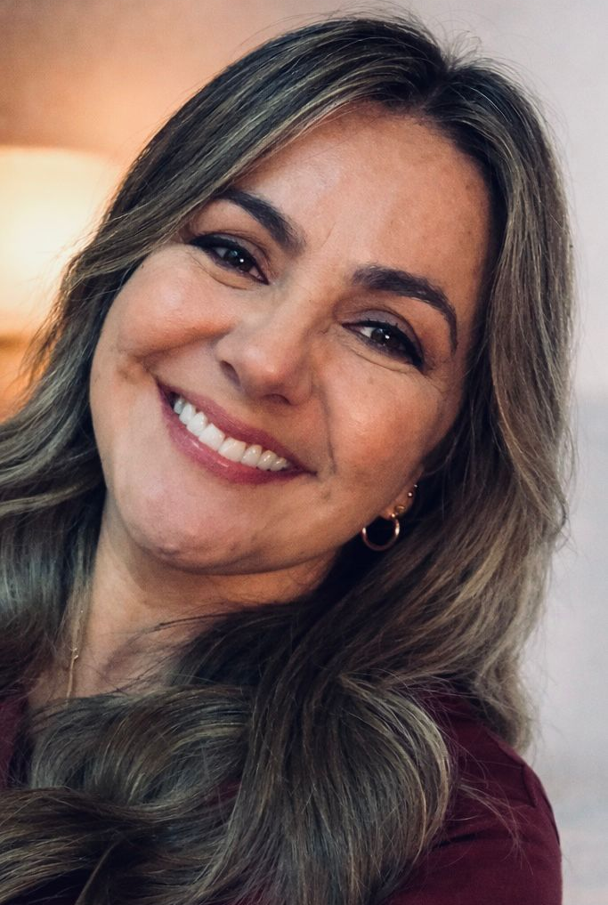 Una mujer con cabello castaño ondulado sonríe a la cámara, luciendo aros y un top color burdeos.