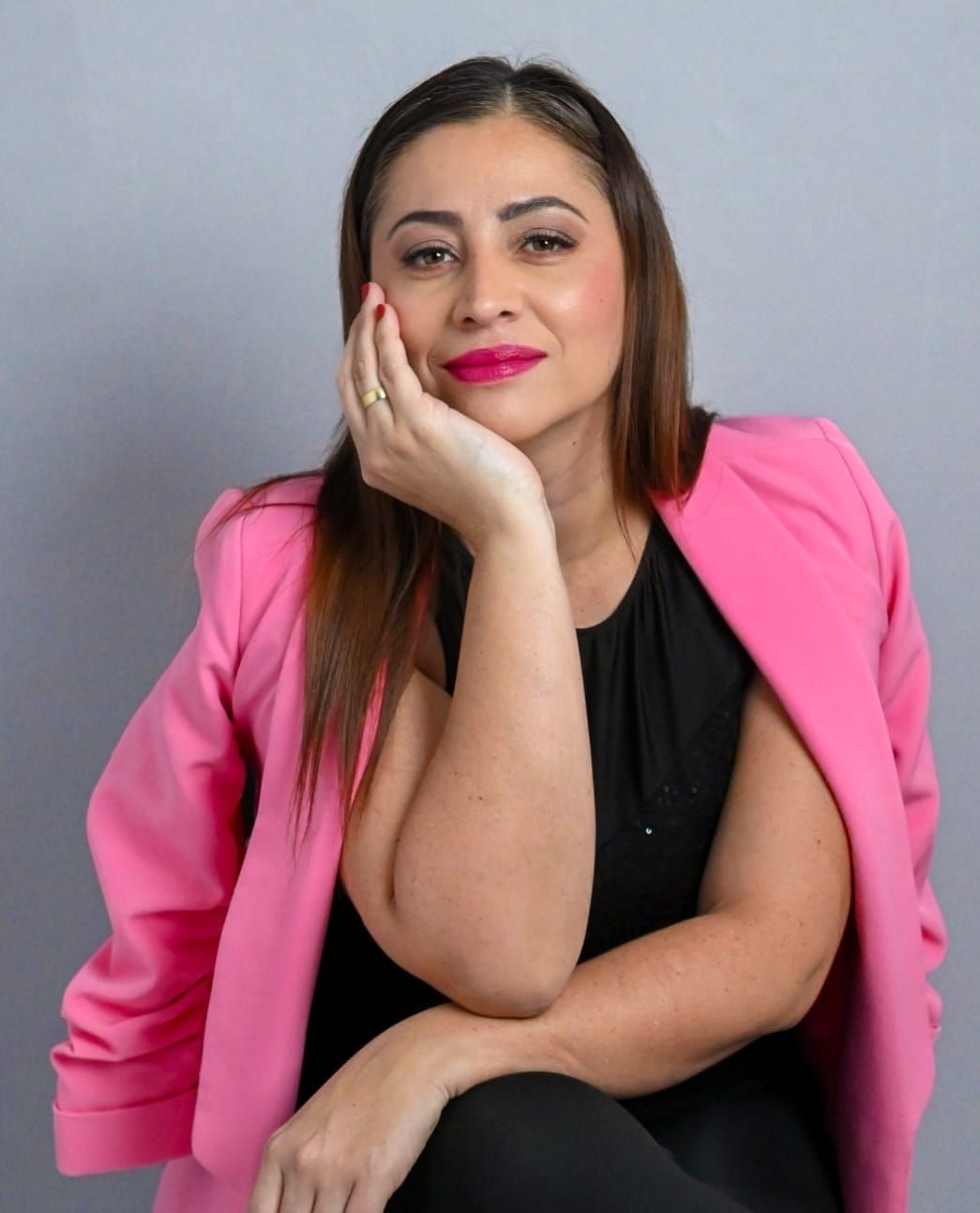 Mujer con blazer rosa, apoyando la barbilla en la mano, mirando a la cámara. Fondo gris.