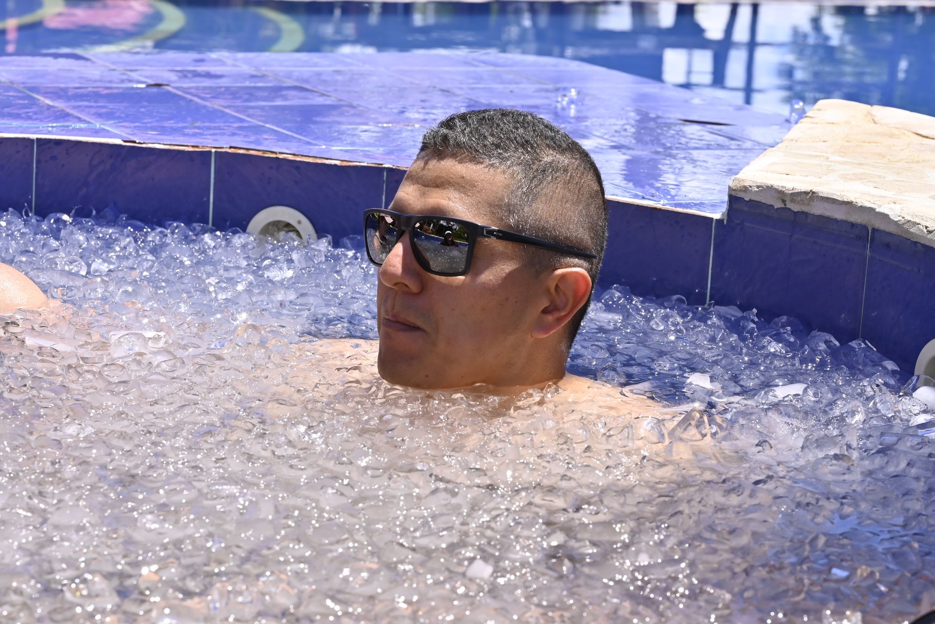 Un hombre que lleva gafas de sol está sentado en un baño de hielo en una bañera con azulejos azules, mirando fuera de cámara.