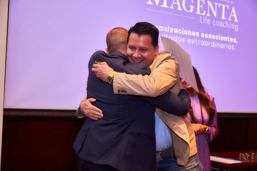 Dos hombres se abrazan en el escenario con una pantalla de fondo. Uno lleva traje y el otro, chaqueta color canela.