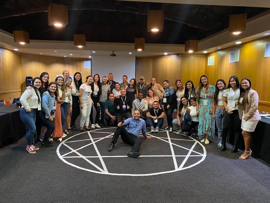 Foto grupal en una sala de conferencias. Gente sonriendo. Un hombre está sentado en el suelo dentro de un símbolo blanco, frente a una pantalla de proyección.