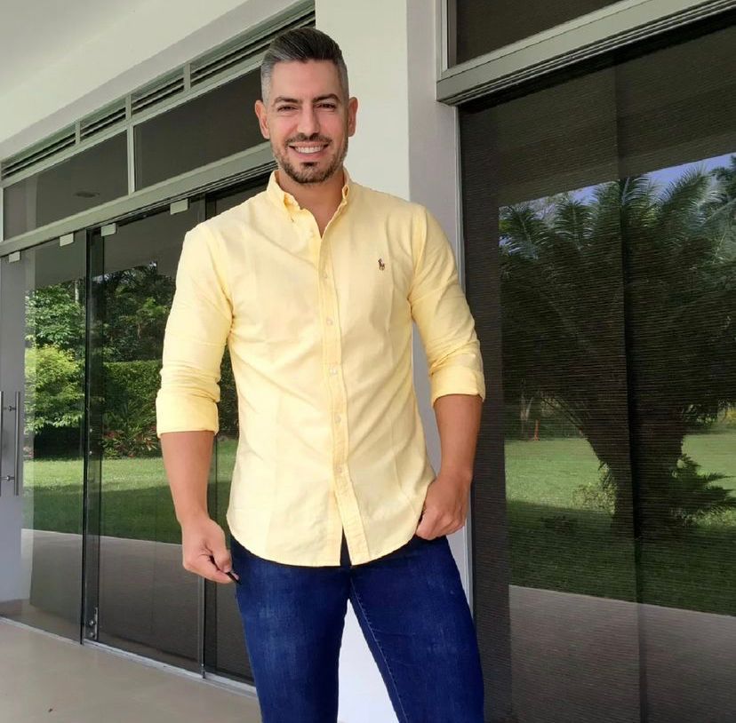 Un hombre con camisa amarilla y jeans sonríe, de pie frente a una puerta de vidrio.