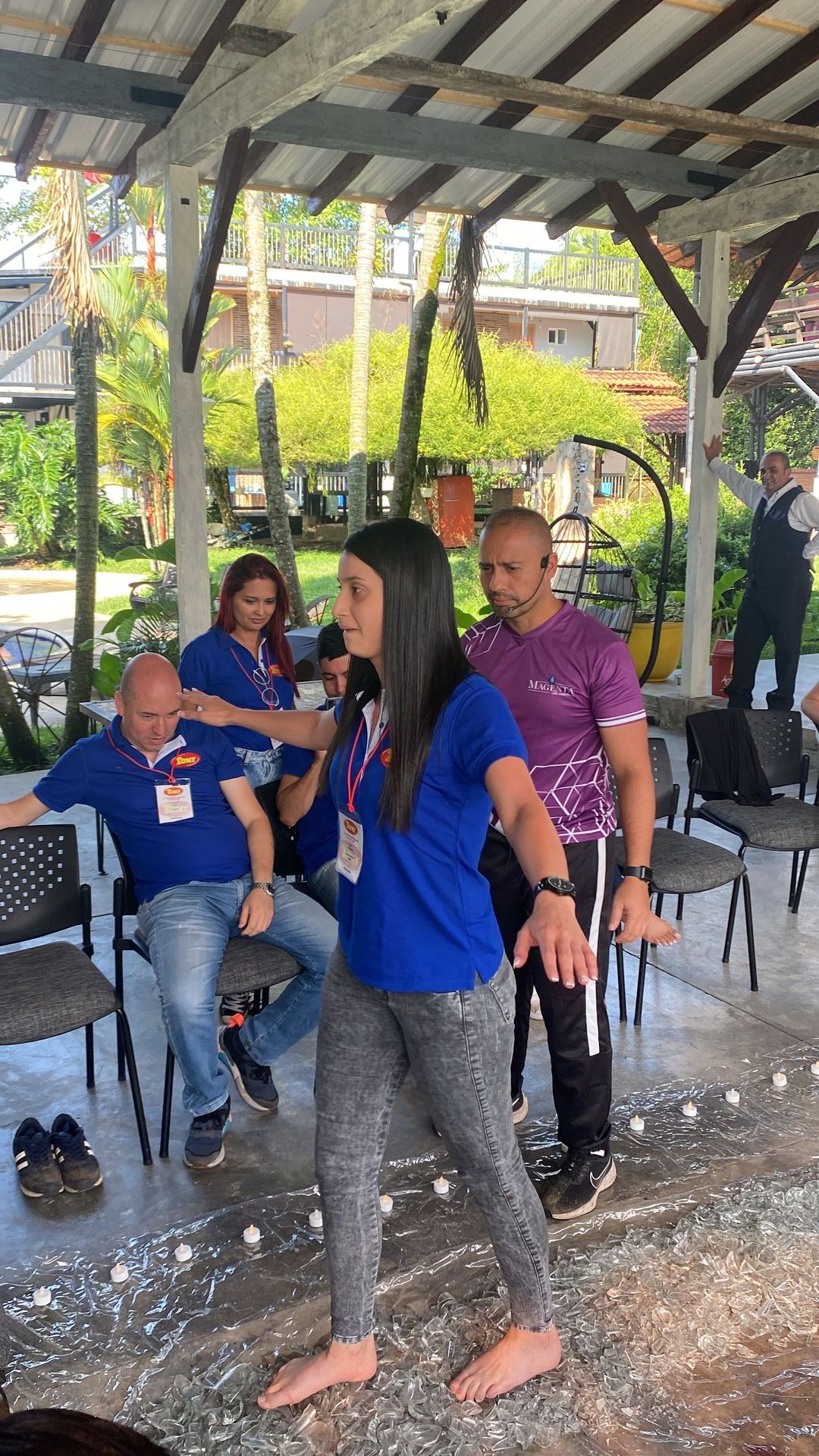 Personas participando en un ejercicio de formación de equipos al aire libre, una mujer haciendo equilibrio descalza, rodeada de otras cerca de sillas.
