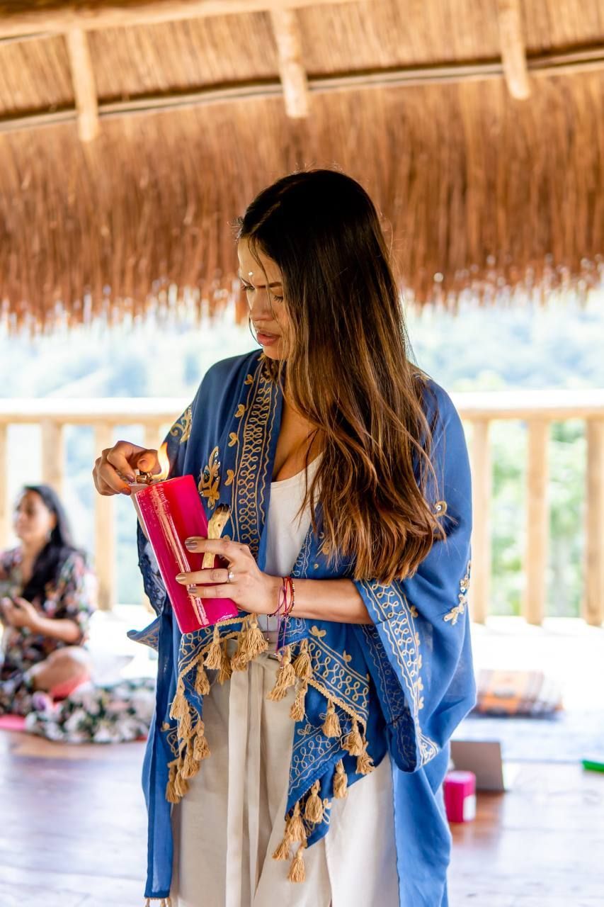 Mujer con un chal azul sosteniendo un objeto rosa, al aire libre en una terraza de madera bajo un techo de paja.