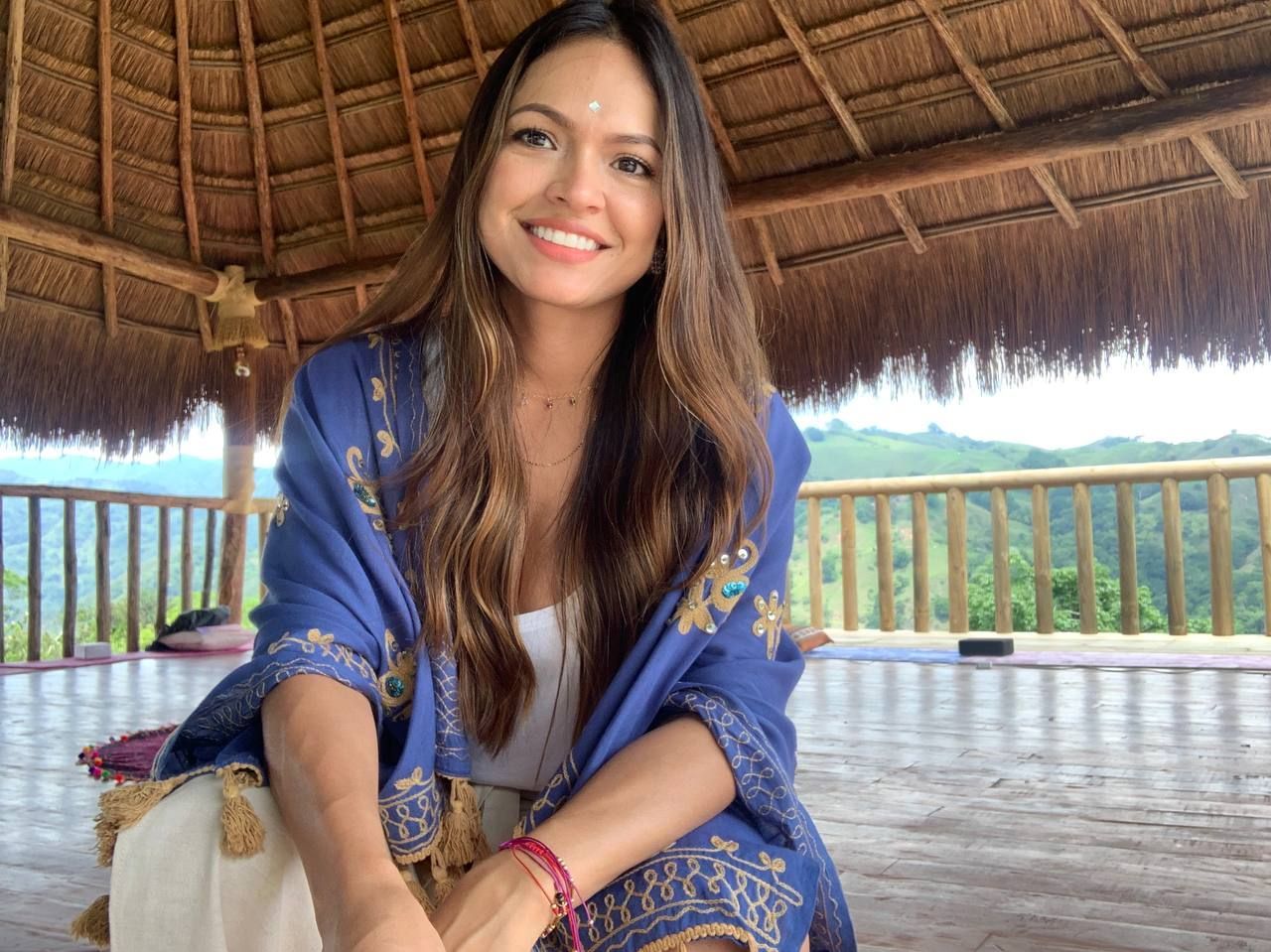 Mujer sonriendo, sentada en un pabellón de madera. Lleva un chal azul y una blusa blanca, mirando a la cámara. Montañas al fondo.