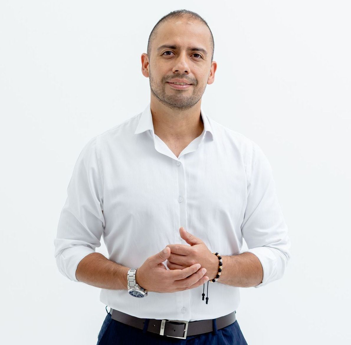 Hombre con camisa blanca, manos entrelazadas, sonriendo, sobre un fondo blanco.