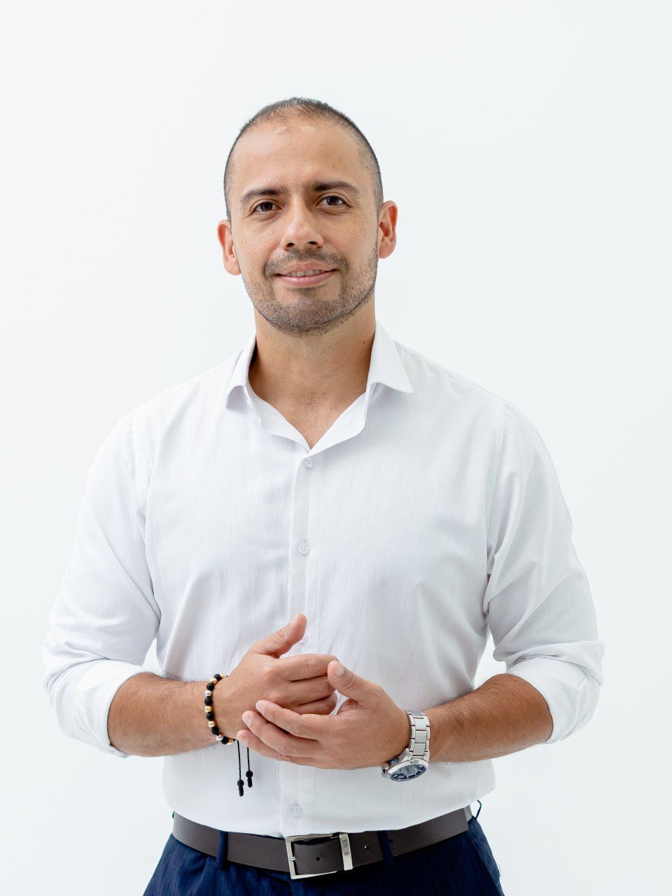 Hombre con camisa blanca, con las manos entrelazadas, sonriendo, sobre fondo blanco.