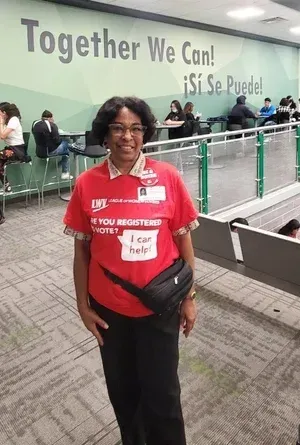 Woman in red shirt and lanyard smiling at a convention center, with “Together We Can! ¡Sí Se Puede!” on the wall.