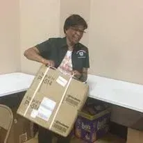 Person in a black shirt holding a large cardboard box in a room with tables and chairs.
