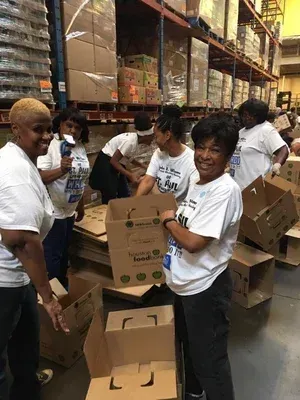Volunteers in a warehouse packing cardboard boxes amid tall shelves of supplies.