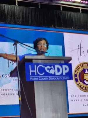Speaker at Harris County Democratic Party podium with blue backdrop and microphone on stage