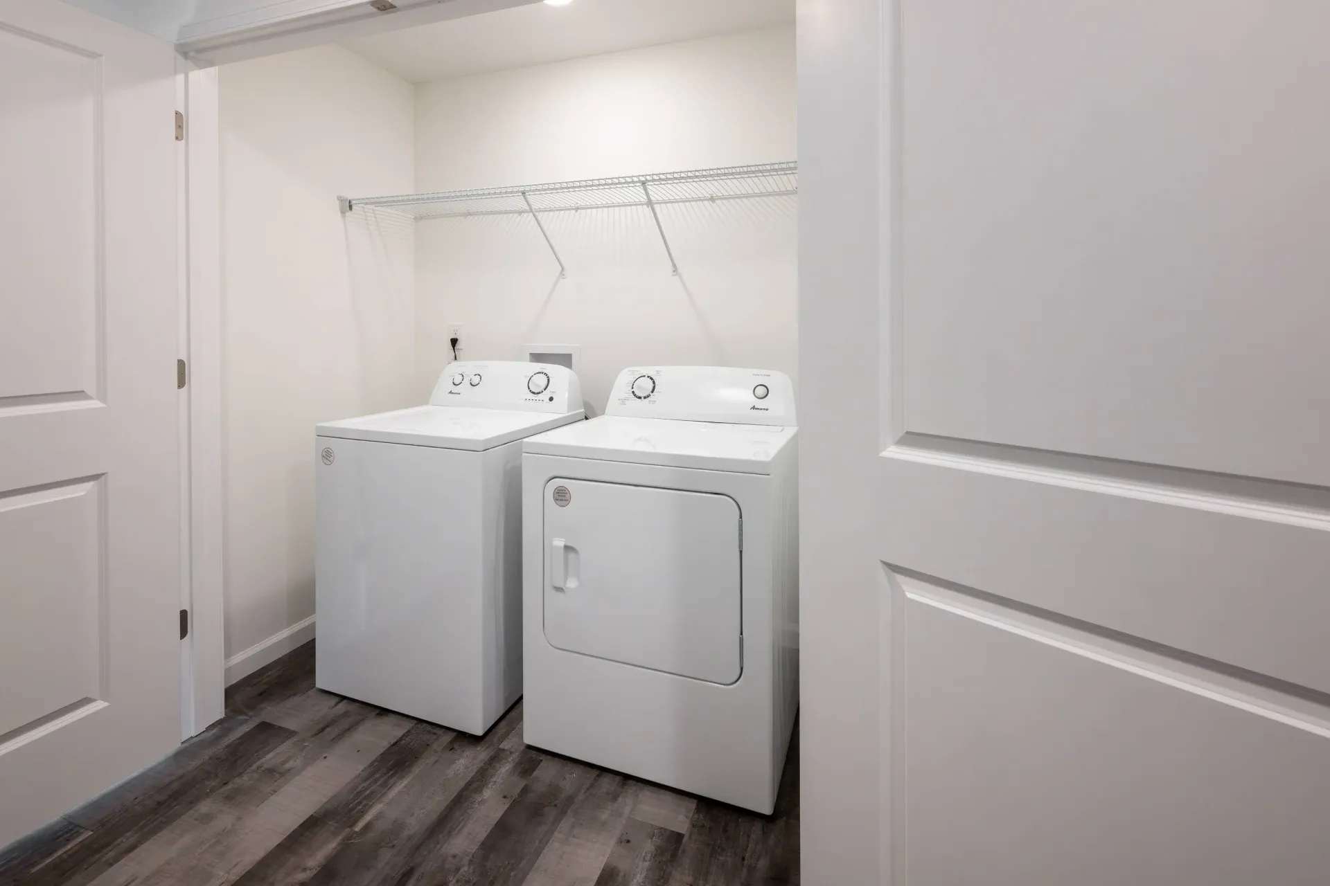 Two white laundry machines (washer and dryer) in a small closet with a wire shelf.
