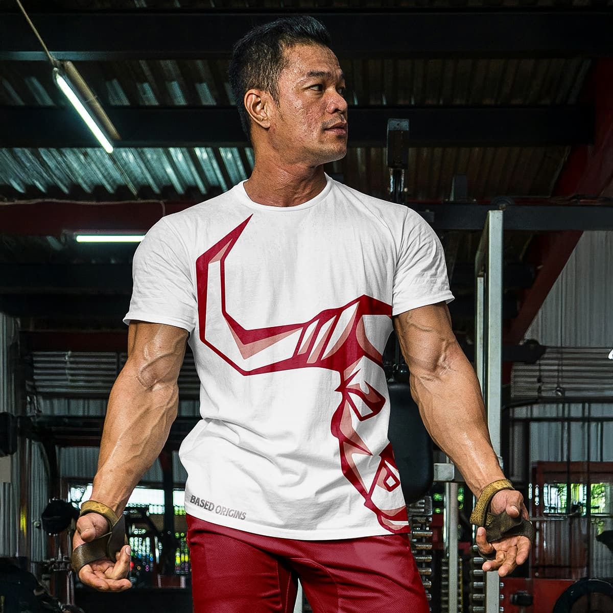 Muscular man in a white graphic T-shirt and red pants standing in a gym, holding dumbbells.