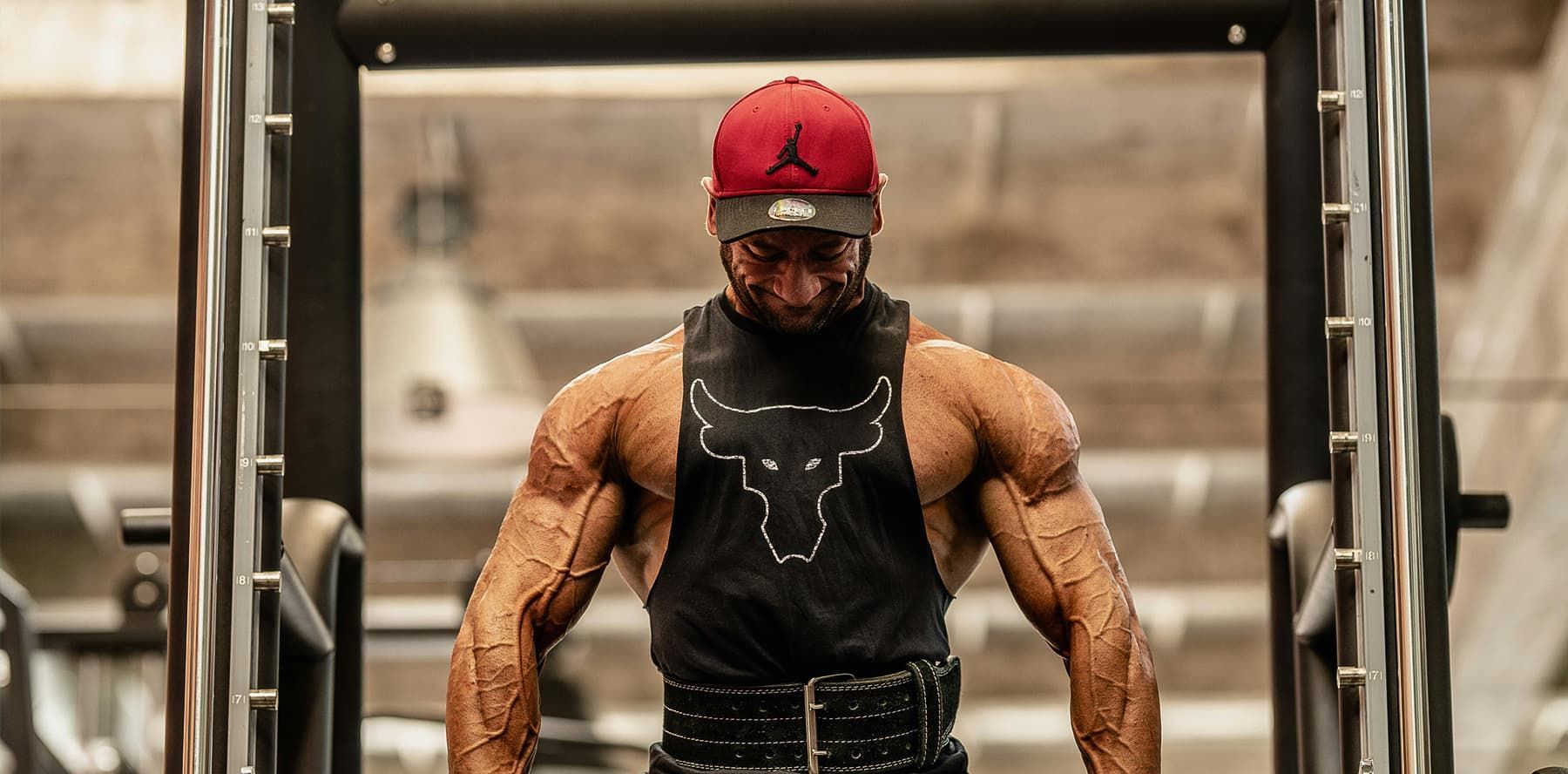 Muscular man in a red cap working out on gym equipment, wearing a black tank top and gloves.