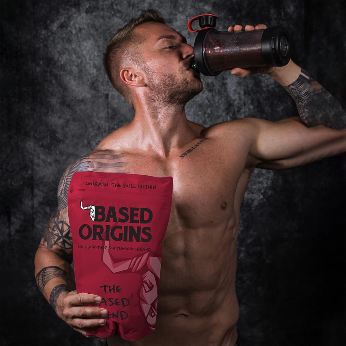 Shirtless man drinking from a shaker and holding a red “Based Origins” bag against a dark backdrop
