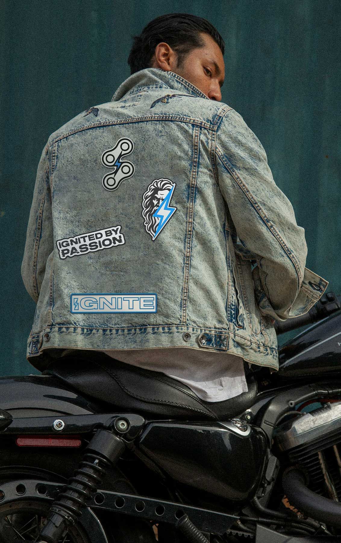 Person sitting on a motorcycle, wearing a faded denim jacket with patches, seen from behind.