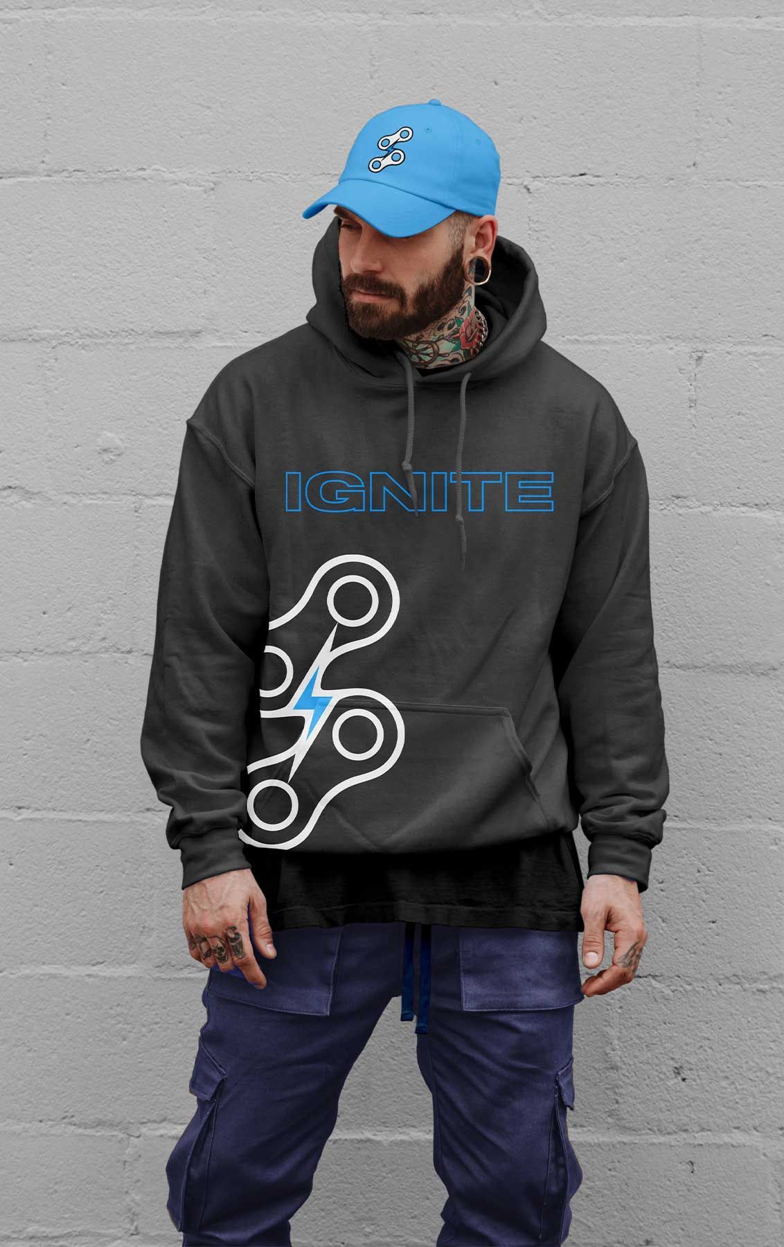Man wearing a blue cap and dark hoodie with graphic design, standing against a gray wall.
