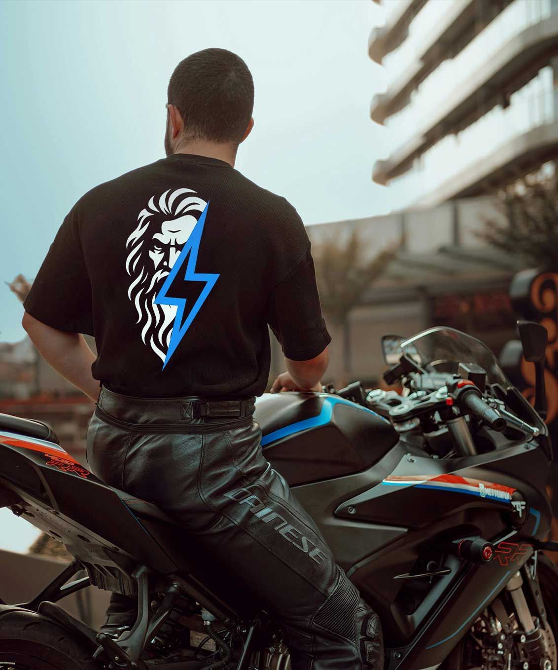 Person in a black T-shirt with a lightning logo stands beside a black-and-blue motorcycle outdoors.