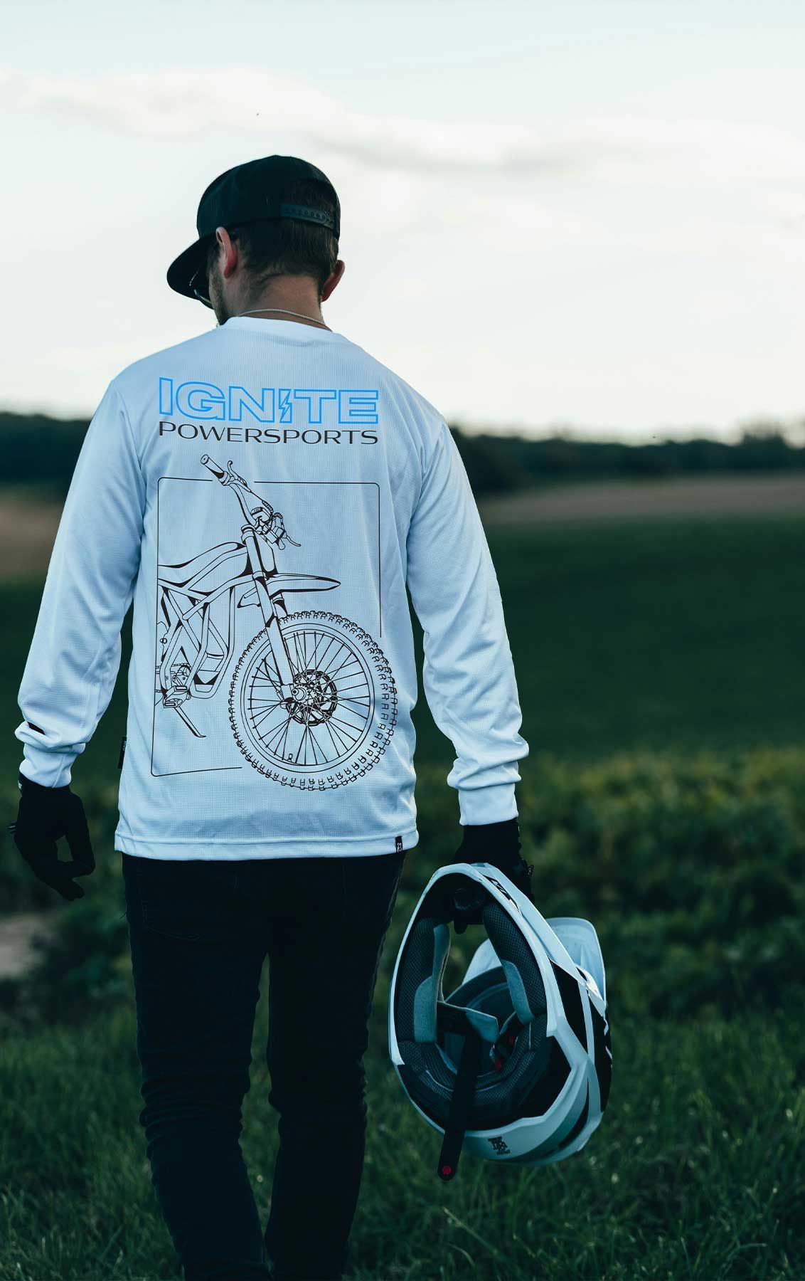 Person in a light gray sweatshirt and black pants stands outdoors holding a backpack at sunset.